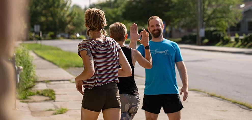 Three people high-five on a sidewalk. The man in a blue shirt smiles. Sunny day.