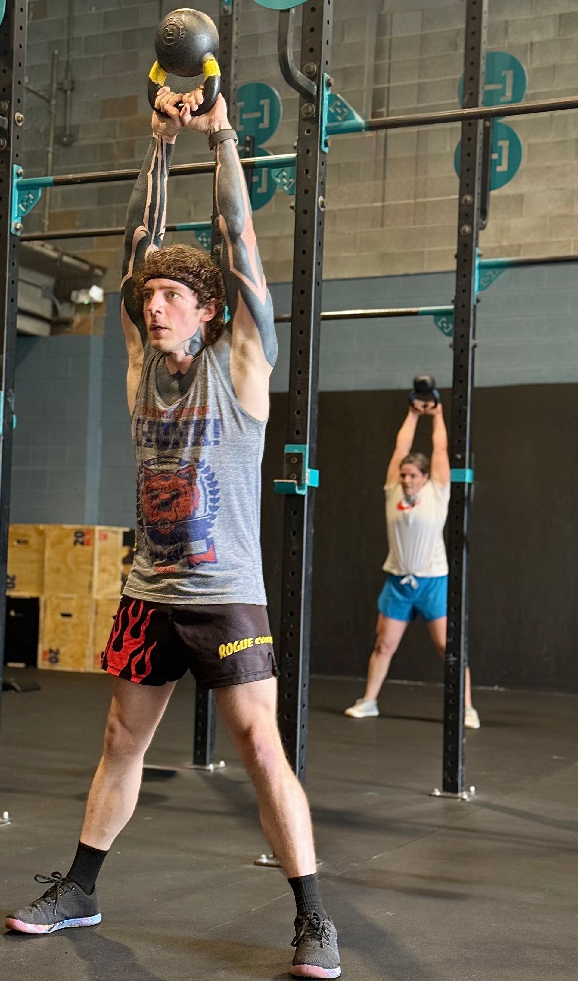 Man in gym in Asheville, NC, lifting kettlebell overhead, other person in background, both wearing activewear.