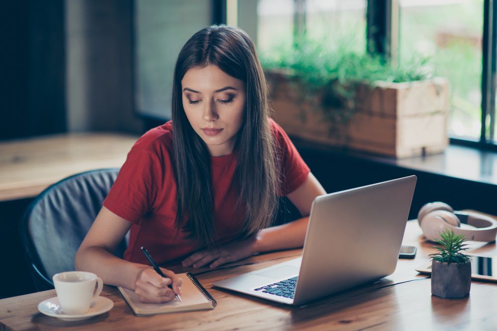 A Woman is Sitting at a Table With a Laptop and Writing in a Notebook — The Molecule Mentor in Lorn, NSW