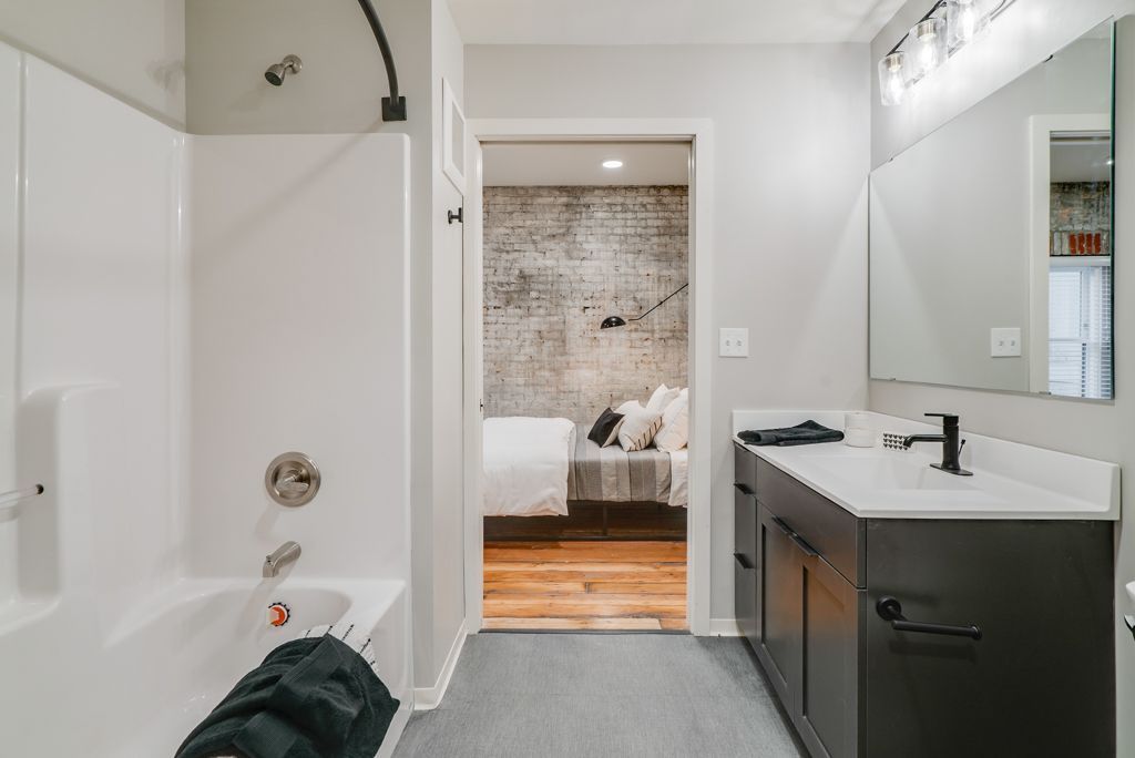 Bathroom with white tub/shower, dark vanity, open door to bedroom with brick wall accent at Commission House, offers apartments in St. Paul, MN.