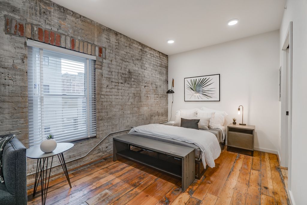 Bedroom with exposed brick wall, window, bed, bench, side table, and wood floor.