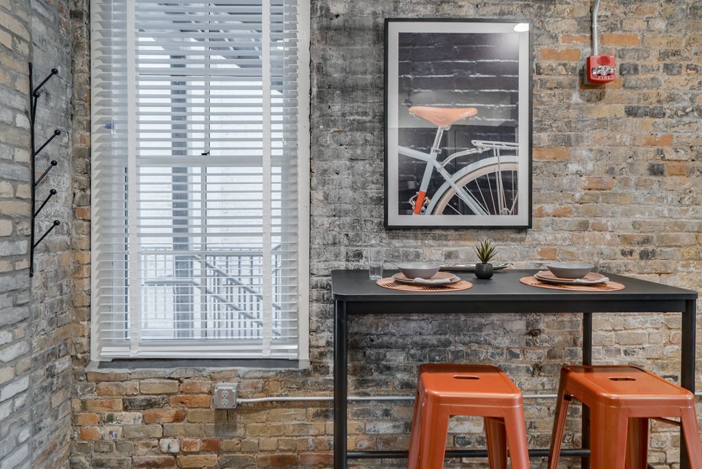 Brick-walled dining area with black table, orange stools, and bicycle art at Commission House, offers apartments in St. Paul, MN.Window with blinds, coat rack.