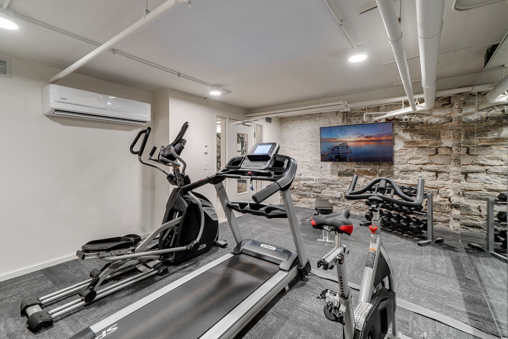 Gym with elliptical, treadmill, stationary bikes, and TV on a stone wall at Commission House, offers apartments in Lowertown St Paul, MN.