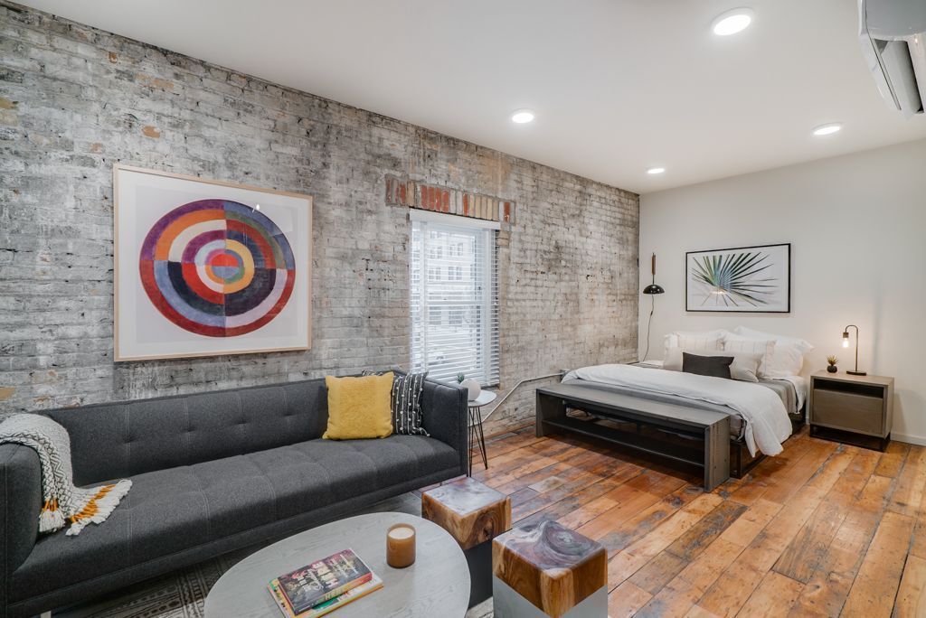 Studio apartment interior with exposed brick wall, gray sofa, bed, and artwork at Commission House, offers apartments in St. Paul, MN.