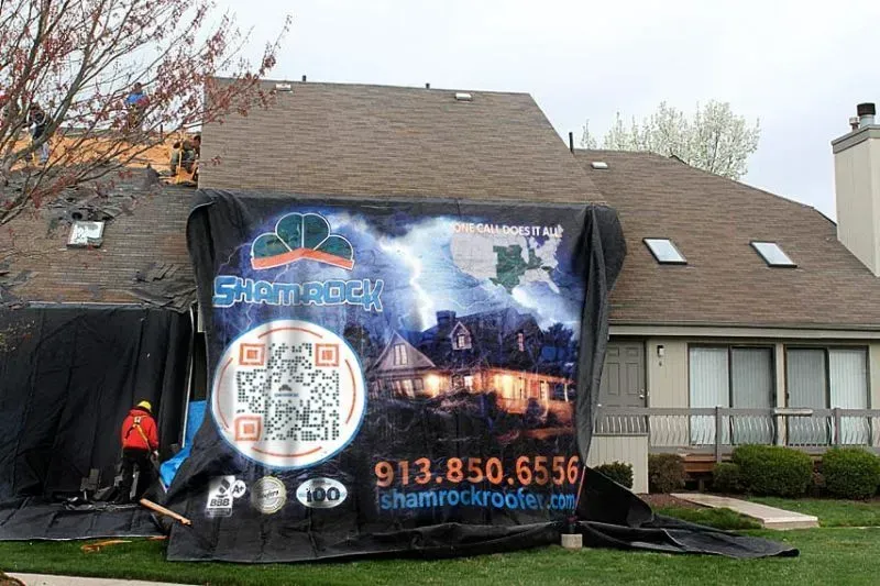 House with roofing work in progress, covered by a black tarp featuring a storm scene and company logo.