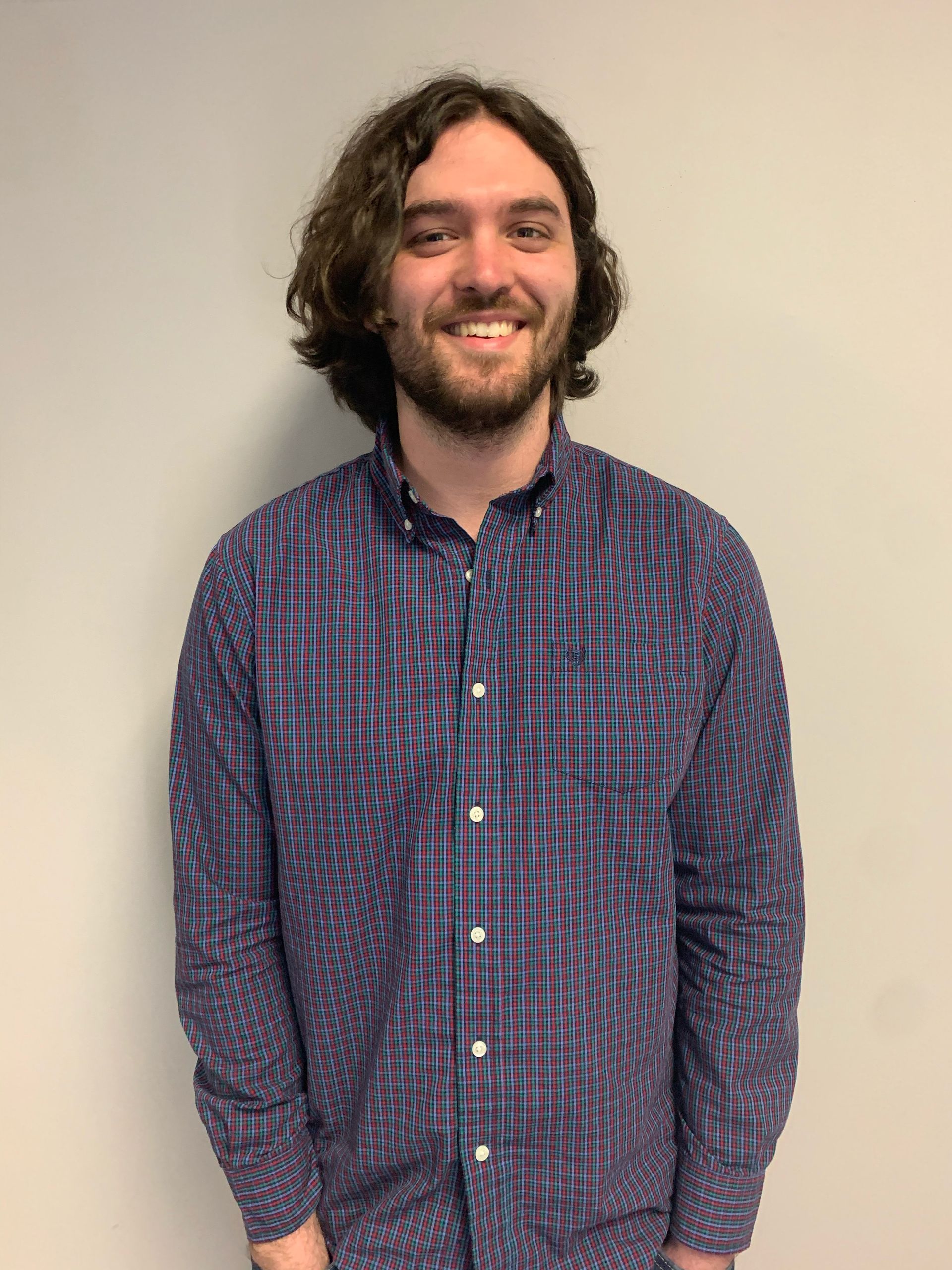 Man with wavy brown hair smiles, wearing a blue and purple plaid shirt, standing in front of a neutral wall.