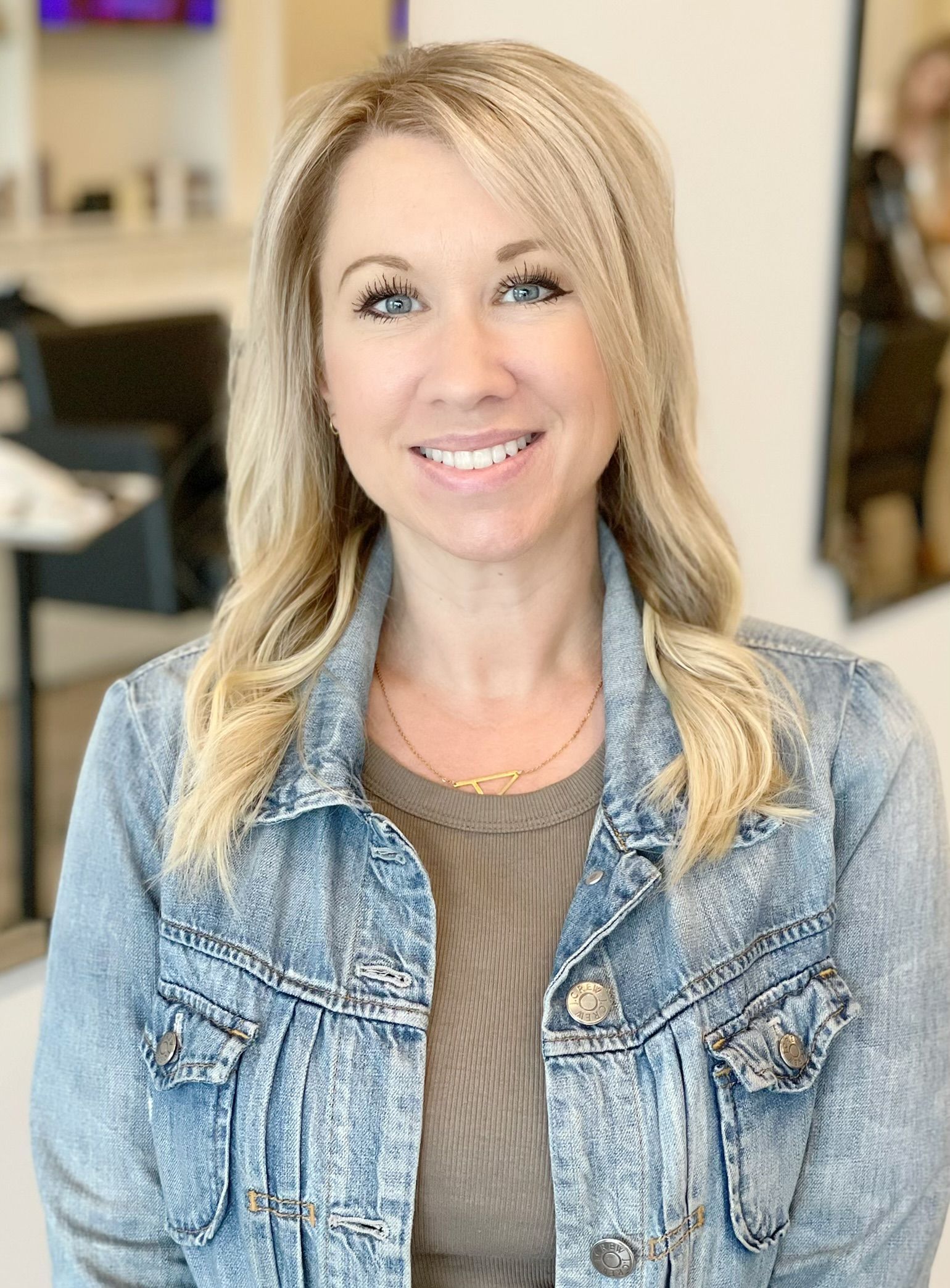 Blonde woman smiles, wearing denim jacket and gold necklace in an office setting.