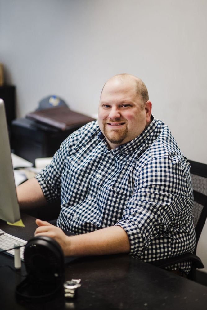 Man in checkered shirt smiles at the camera while sitting at a desk with a computer and headphones.