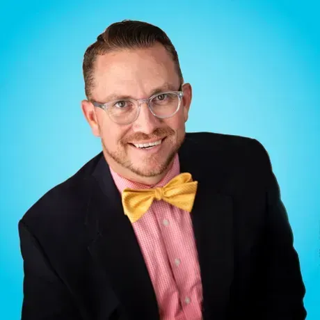 Man in glasses, yellow bow tie, navy blazer, pink shirt, smiling against a blue background.