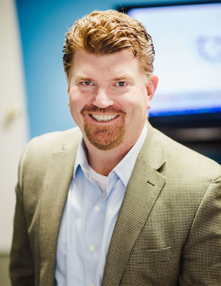 Man with reddish hair, smiling, wearing a light blue shirt and tan blazer, in front of a blue wall and a TV screen.