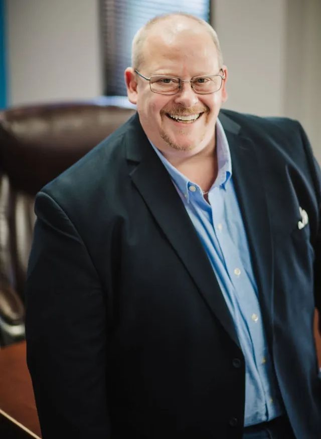 Man smiling, wearing glasses, blue shirt, dark blazer, office setting.