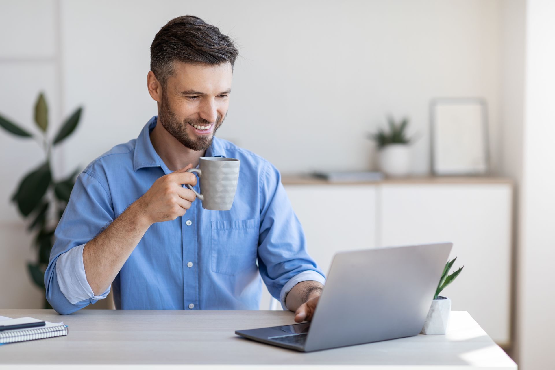 Man smiling, holding a mug, using a laptop at a desk indoors.