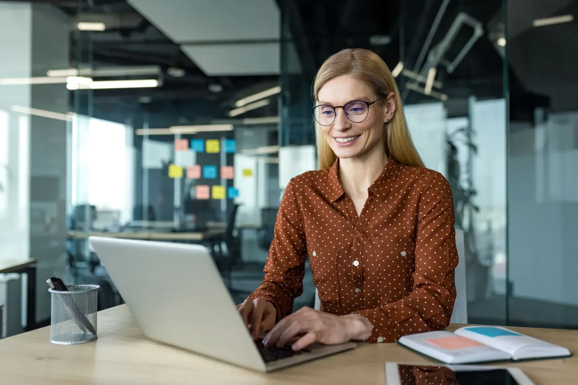 Woman with blonde hair wearing glasses and brown shirt smiles while working on laptop in modern office.
