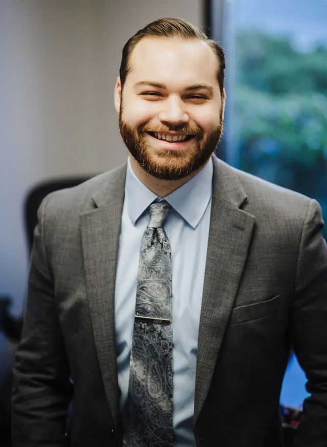 Man in suit smiles at the camera, tie and light blue shirt. Indoor setting.