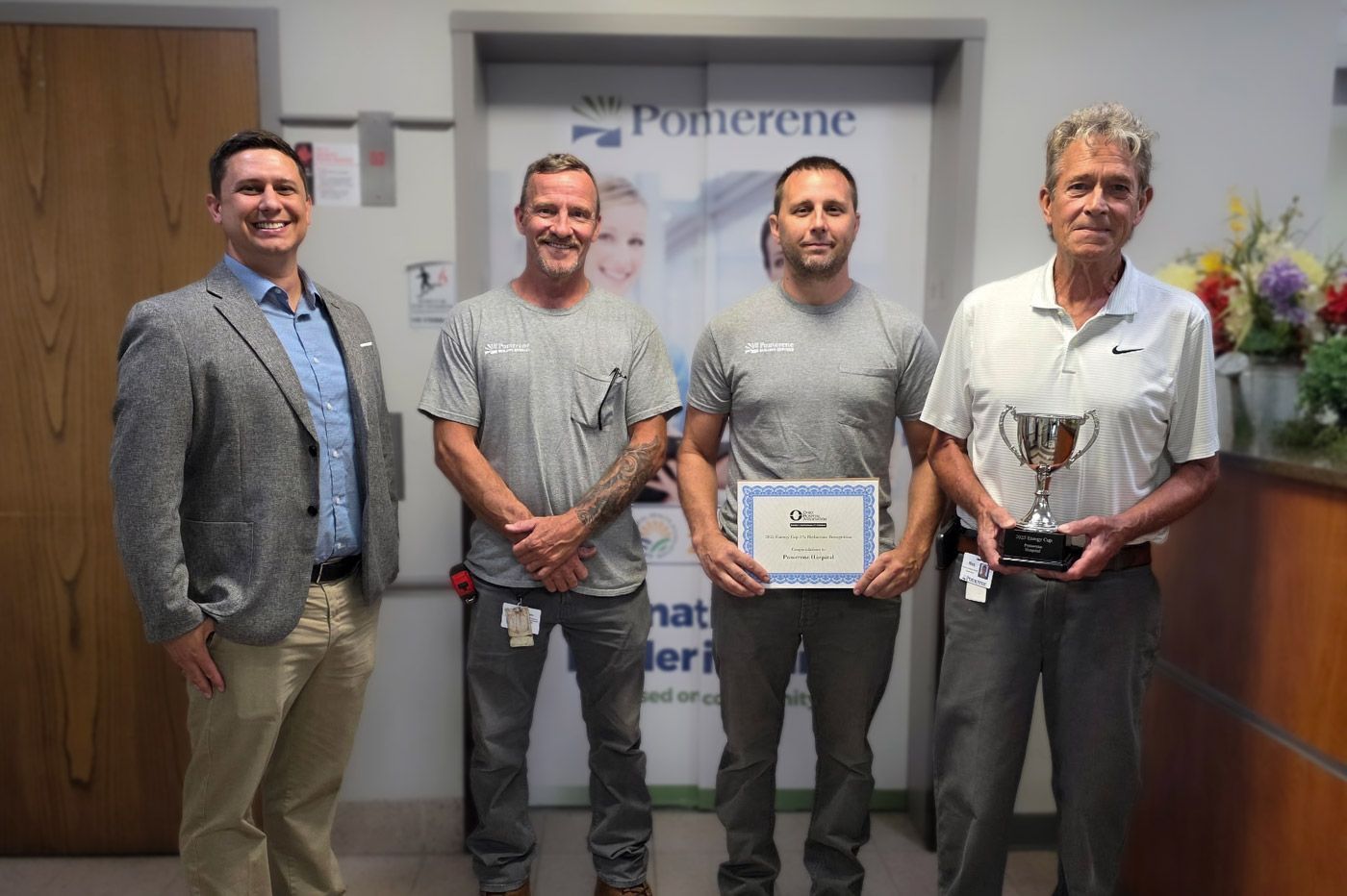 Four men in a well-lit office. Two men hold a certificate, and one holds a trophy. The background has a Pomerene sign.