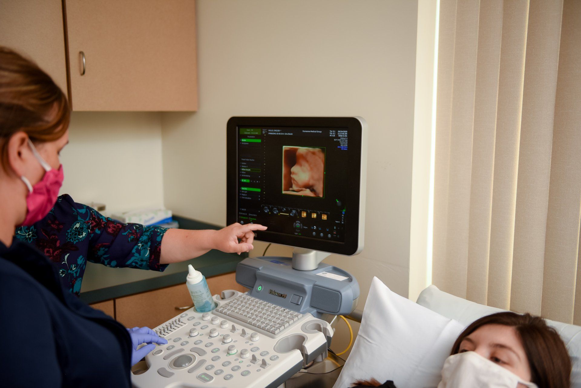 Nurse points to ultrasound image of a fetus. Patient rests, both wearing masks. Examination room.