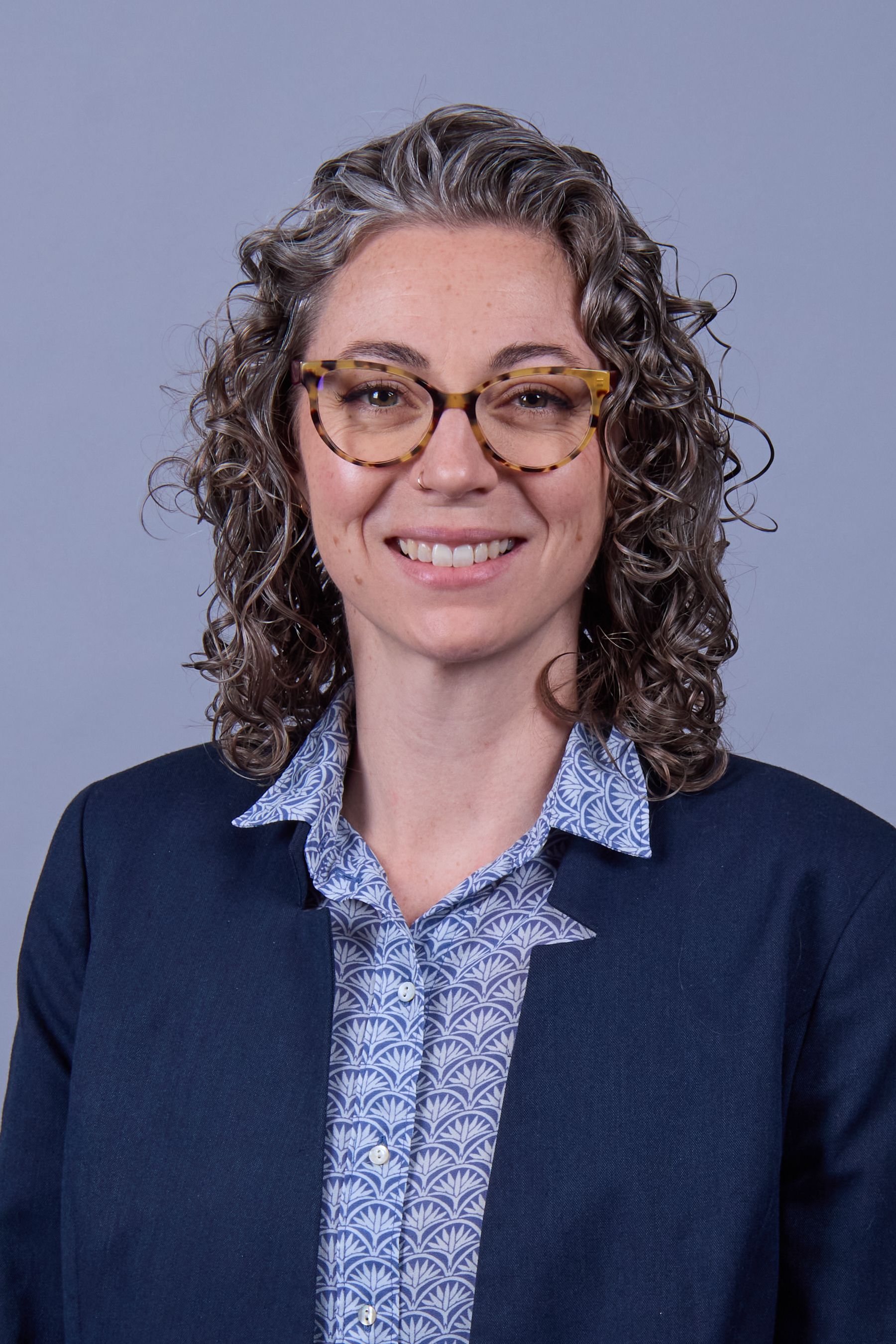 Woman with curly gray hair and glasses, wearing a blue blazer and patterned button-down shirt, smiling at the camera.