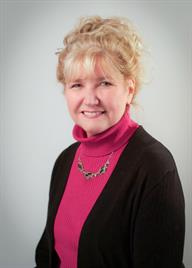 Woman with blonde curly hair smiles, wearing a pink turtleneck, black cardigan.
