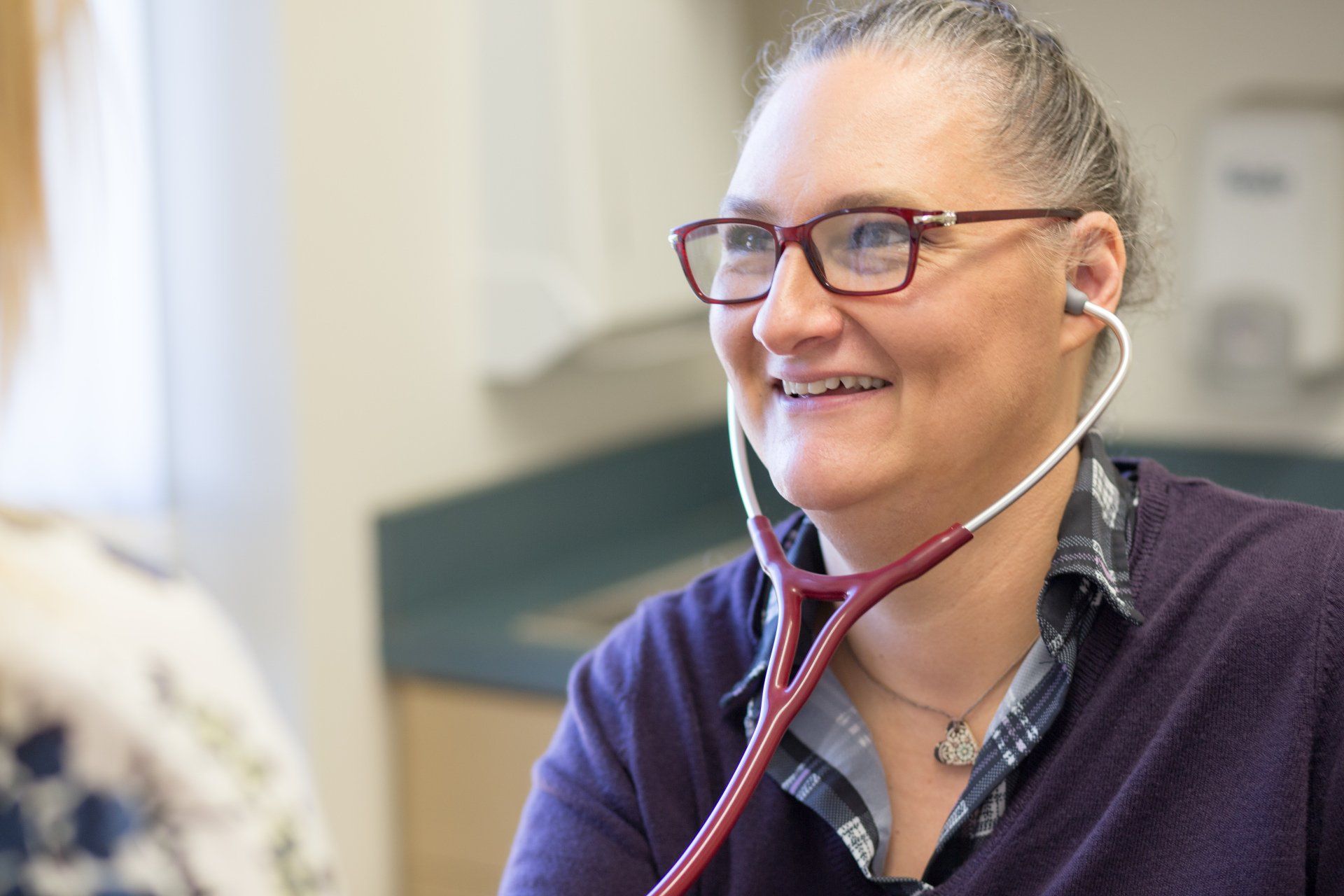 Doctor with stethoscope smiling at a patient; wearing glasses and a purple sweater, in an examination room.