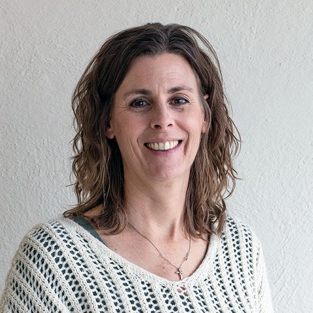 Woman with brown hair smiles at the camera, wearing a white sweater and a necklace. White wall in the background.