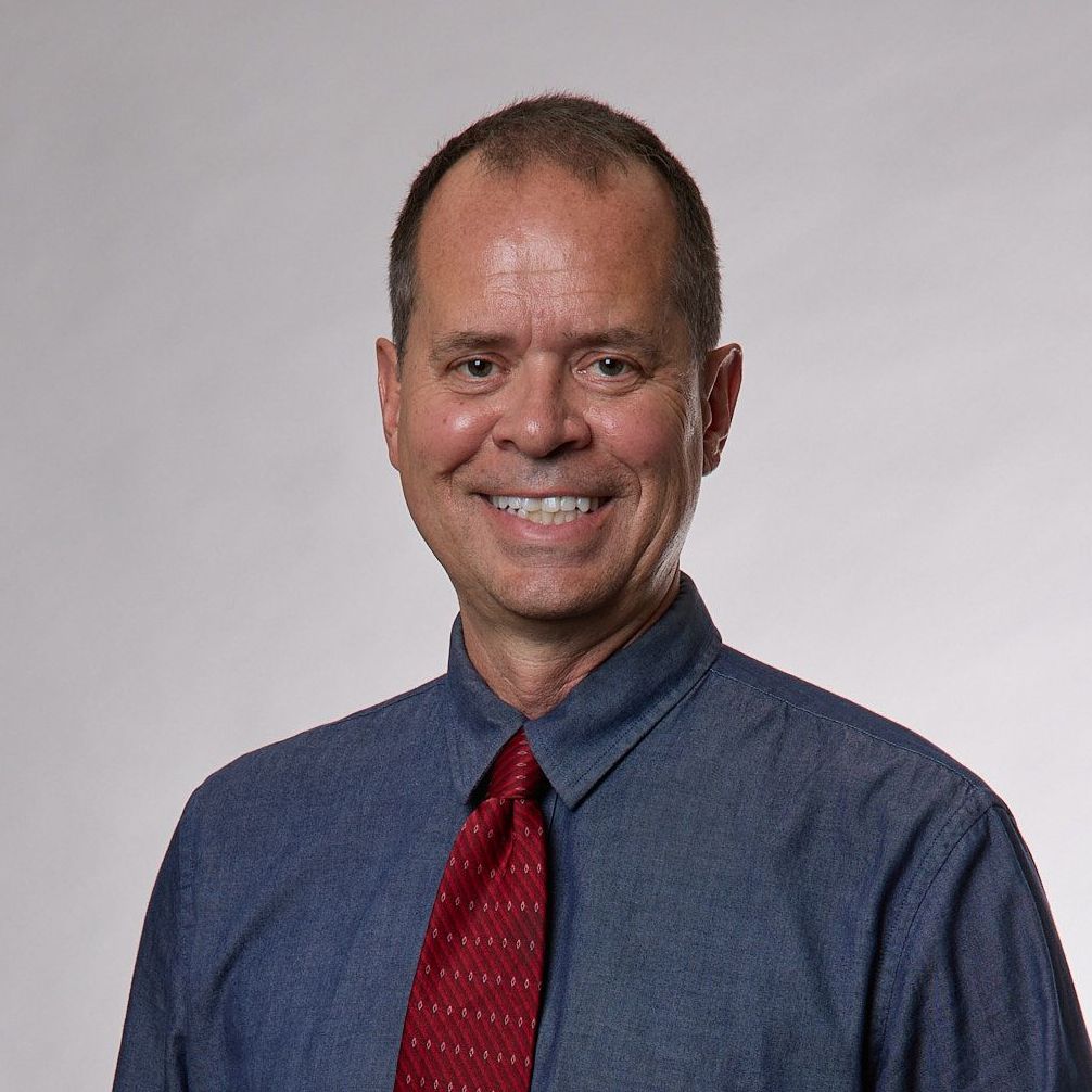 A smiling man with thinning hair wearing a blue shirt and a red tie, against a gray gradient backdrop.