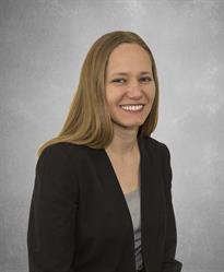 Blonde woman in a black blazer smiles at the camera against a gray background.