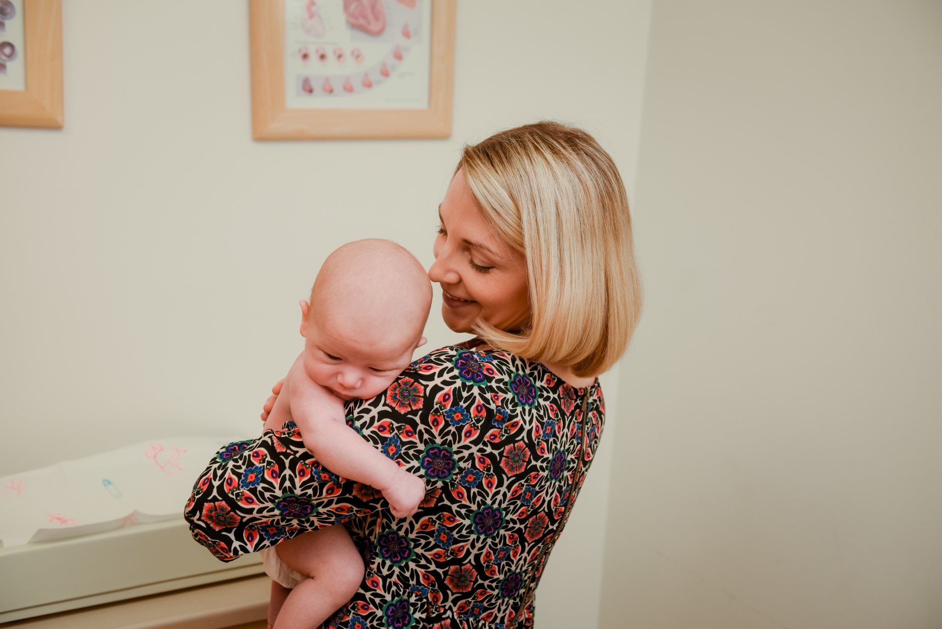 Woman with blonde hair holds a newborn, smiling down at the baby in a room with framed pictures.