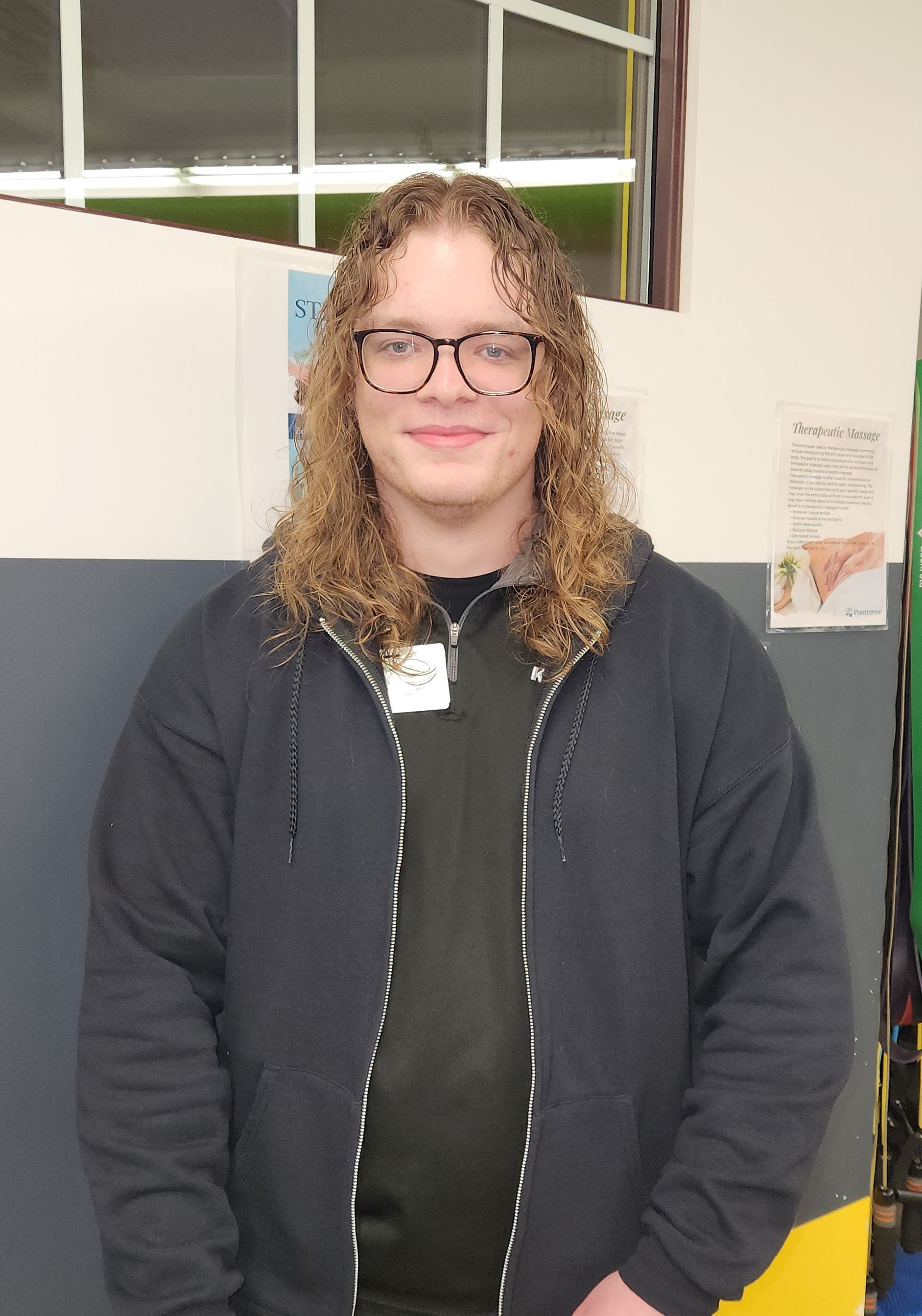 A young person with curly hair and glasses in a black zip-up hoodie smiles in a school setting.