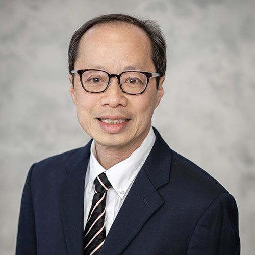 A smiling Asian man wearing glasses, a suit, and a striped tie poses for a headshot against a gray background.