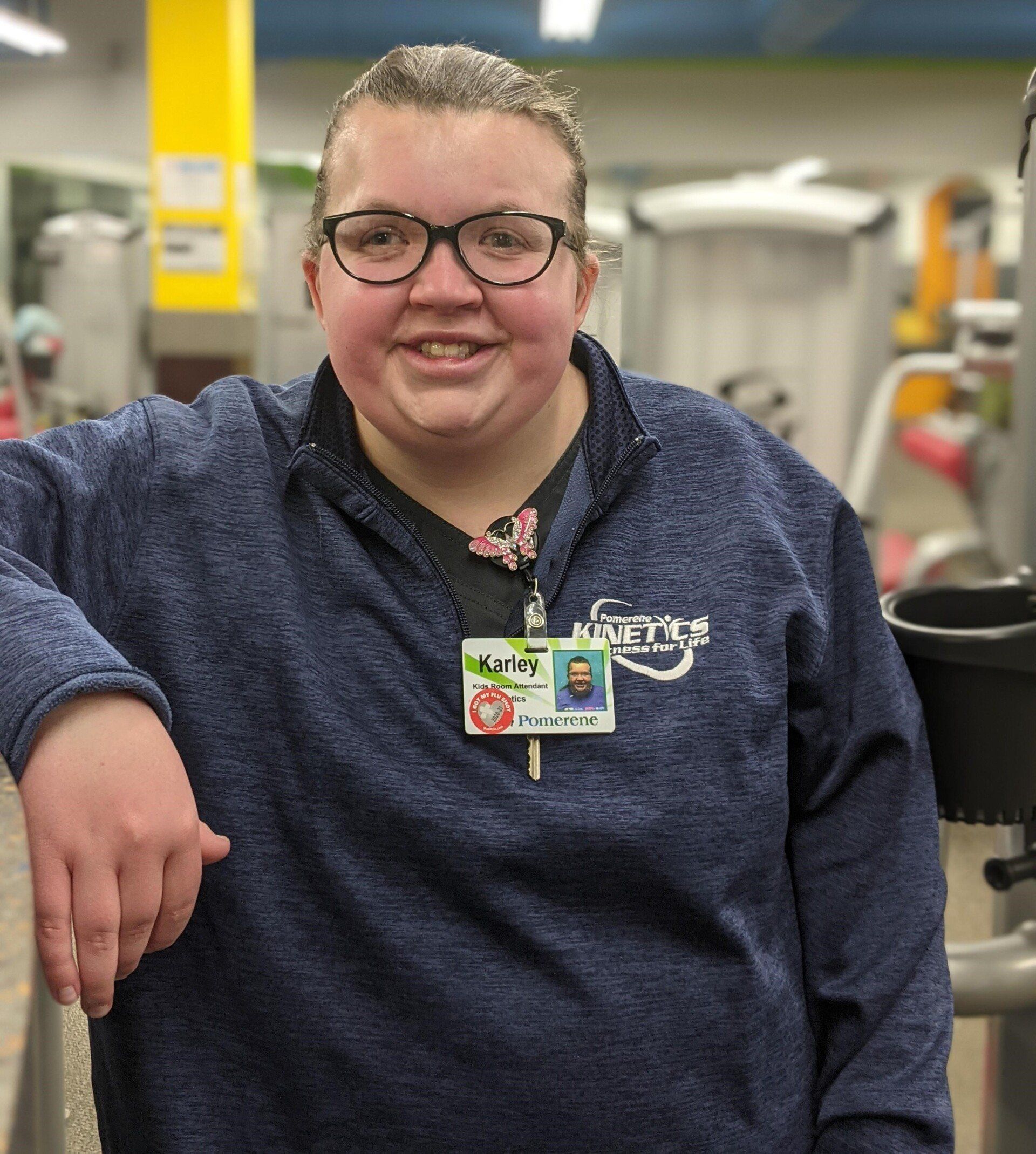 Woman in glasses smiles, leans on a gym machine. She wears a blue sweater and ID badge, gym in background.