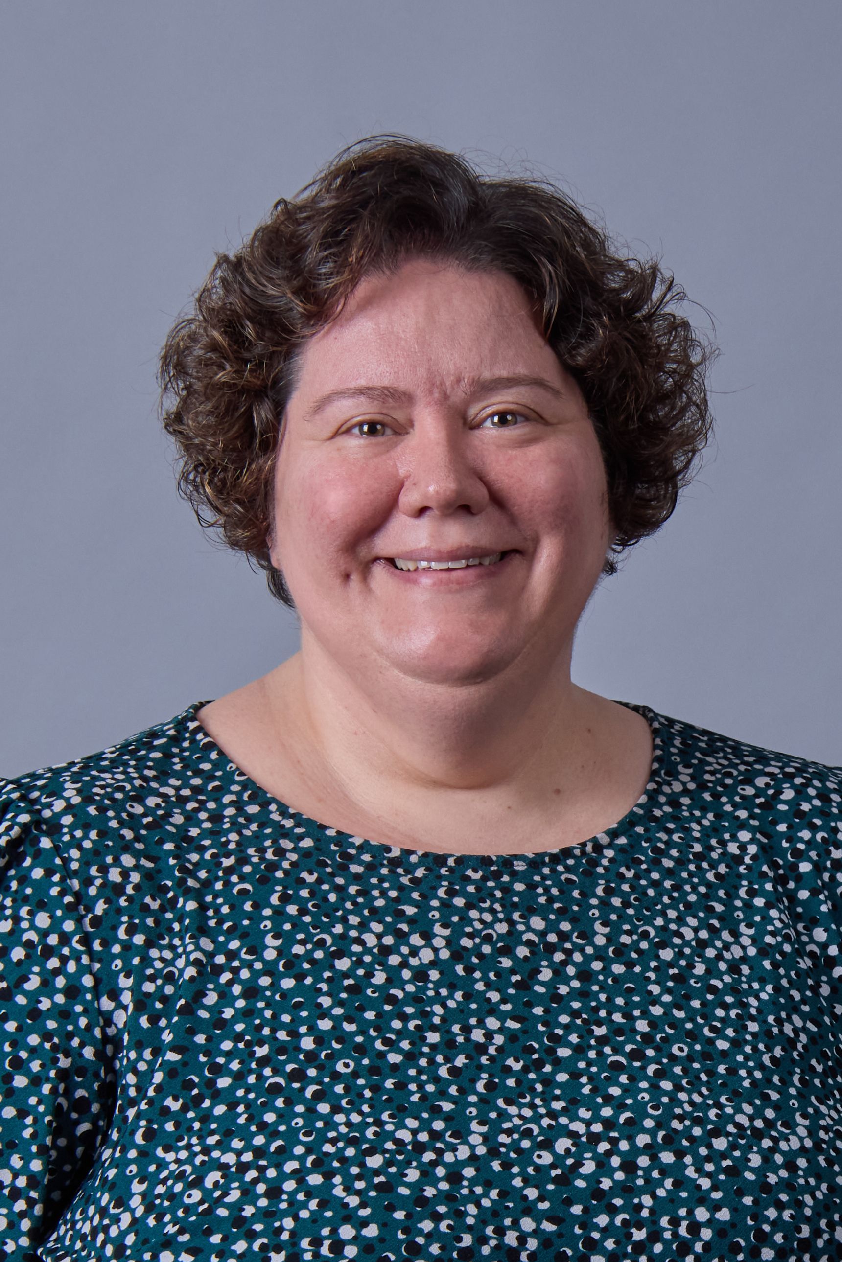 Woman with short, curly brown hair smiles, wearing a teal top with white patterns, set against a gray background.