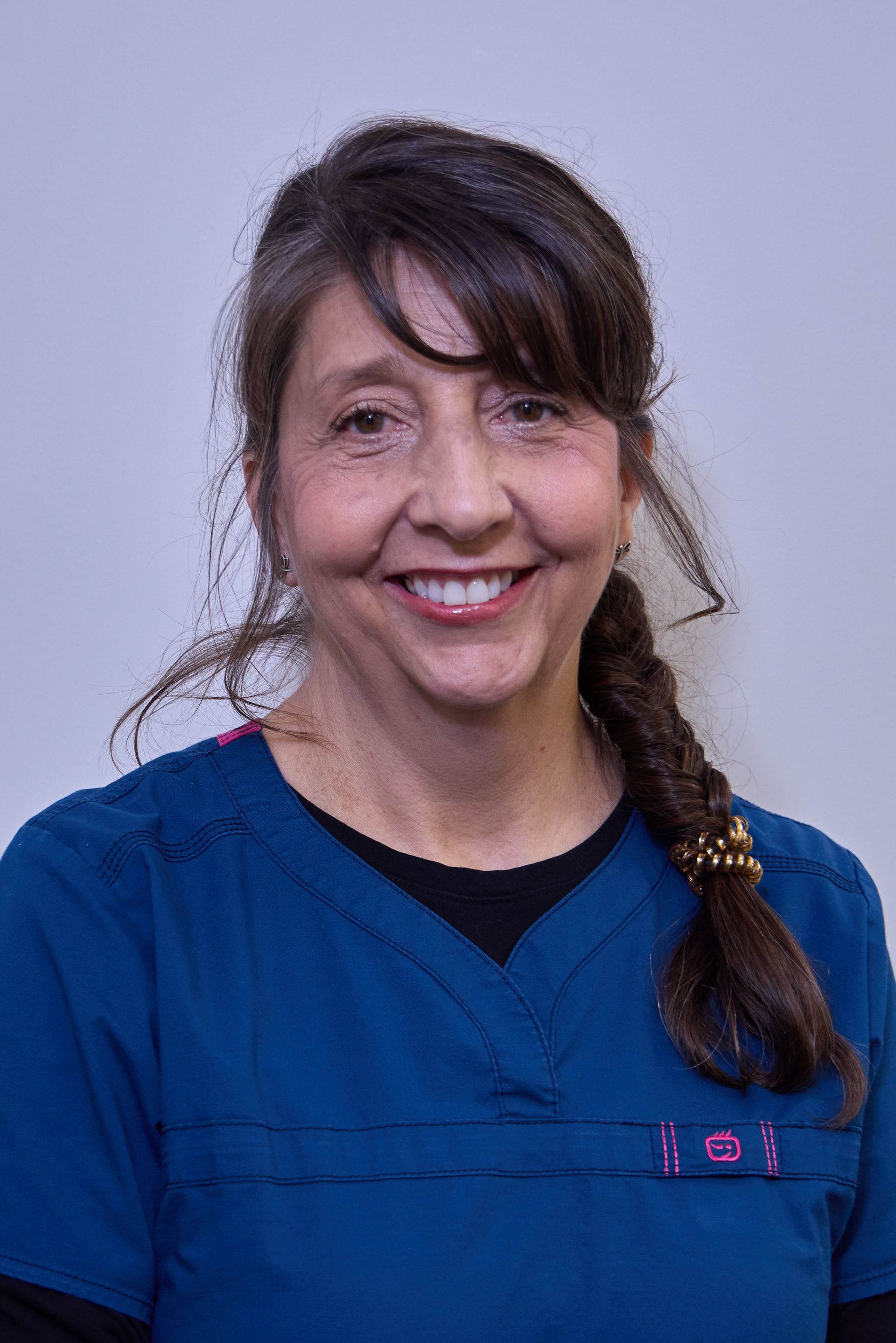 Woman with brown hair in a braid, wearing blue scrubs, smiling in front of a plain white wall.