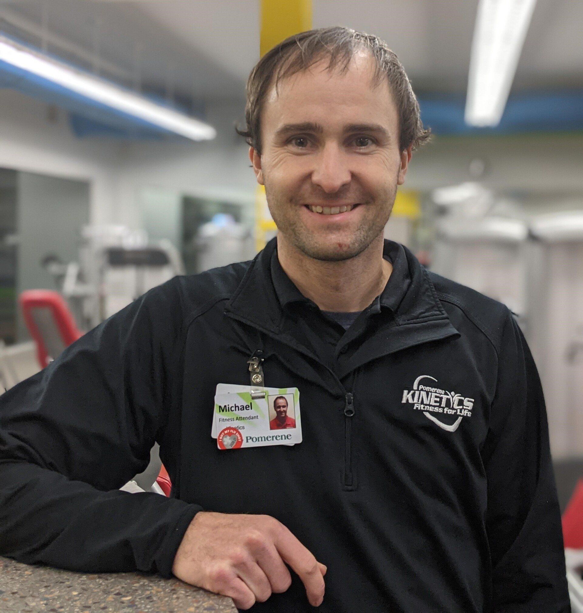 Man in a black jacket with a name tag, smiling in a gym setting.