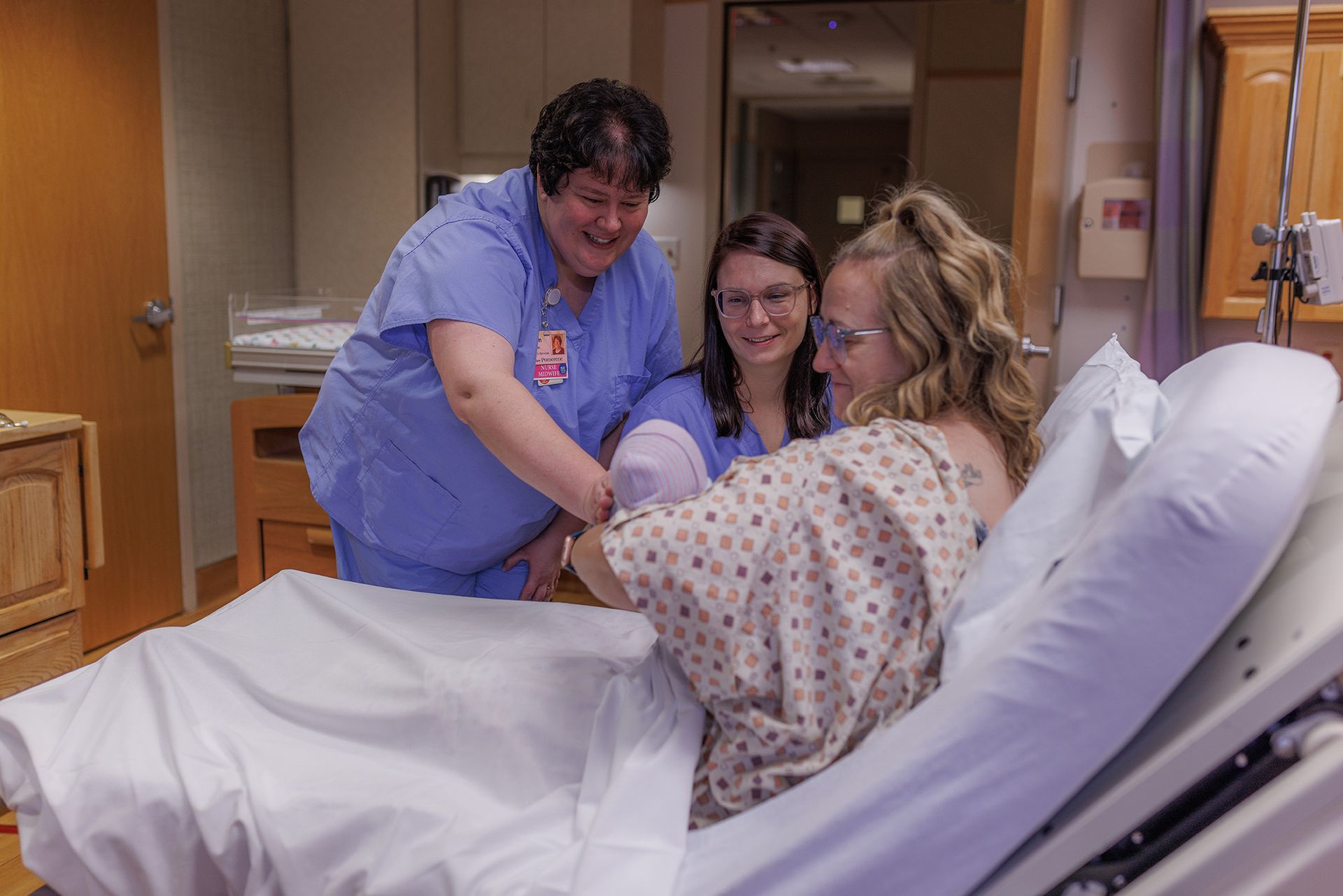 A woman in a hospital bed smiles as nurses help her hold a newborn baby. They are in a light-filled room.