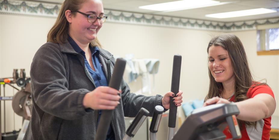 Two women in a light-filled room; one on an elliptical machine, and one assisting. They are smiling.