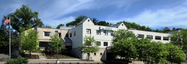 White building with multiple stories under a blue sky. American flag flies to the left, green trees in the foreground.