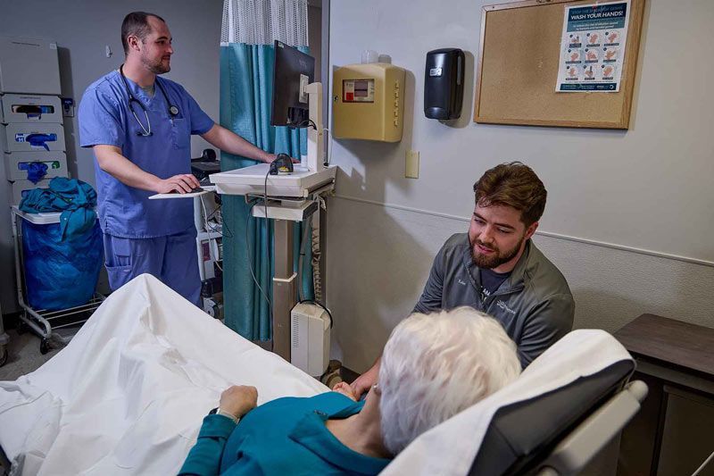 A healthcare worker in scrubs stands by a computer while another speaks to an elderly patient lying in a hospital bed.