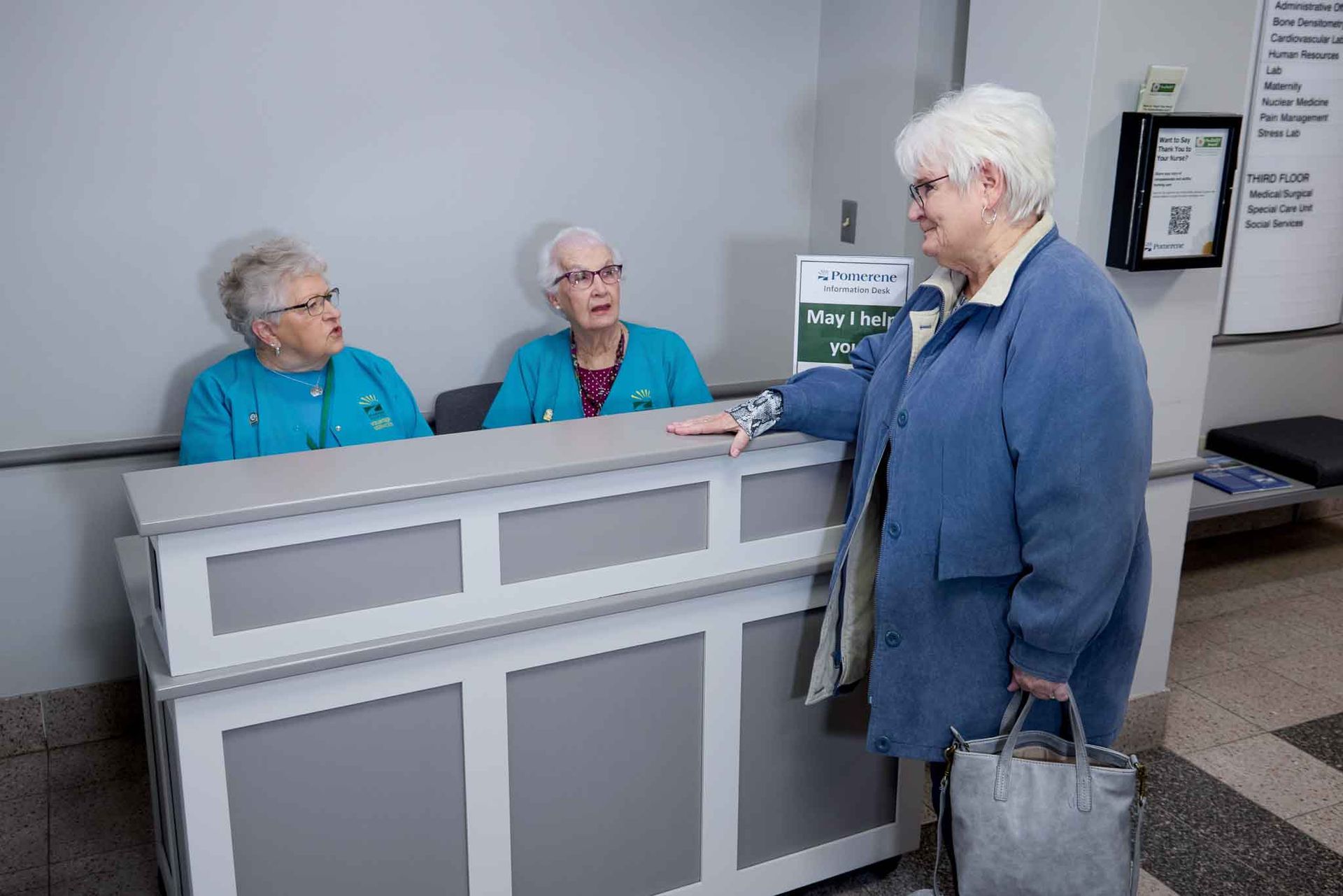 Three older women at a reception desk, two behind it in teal tops, one in a blue jacket standing at the desk, talking.