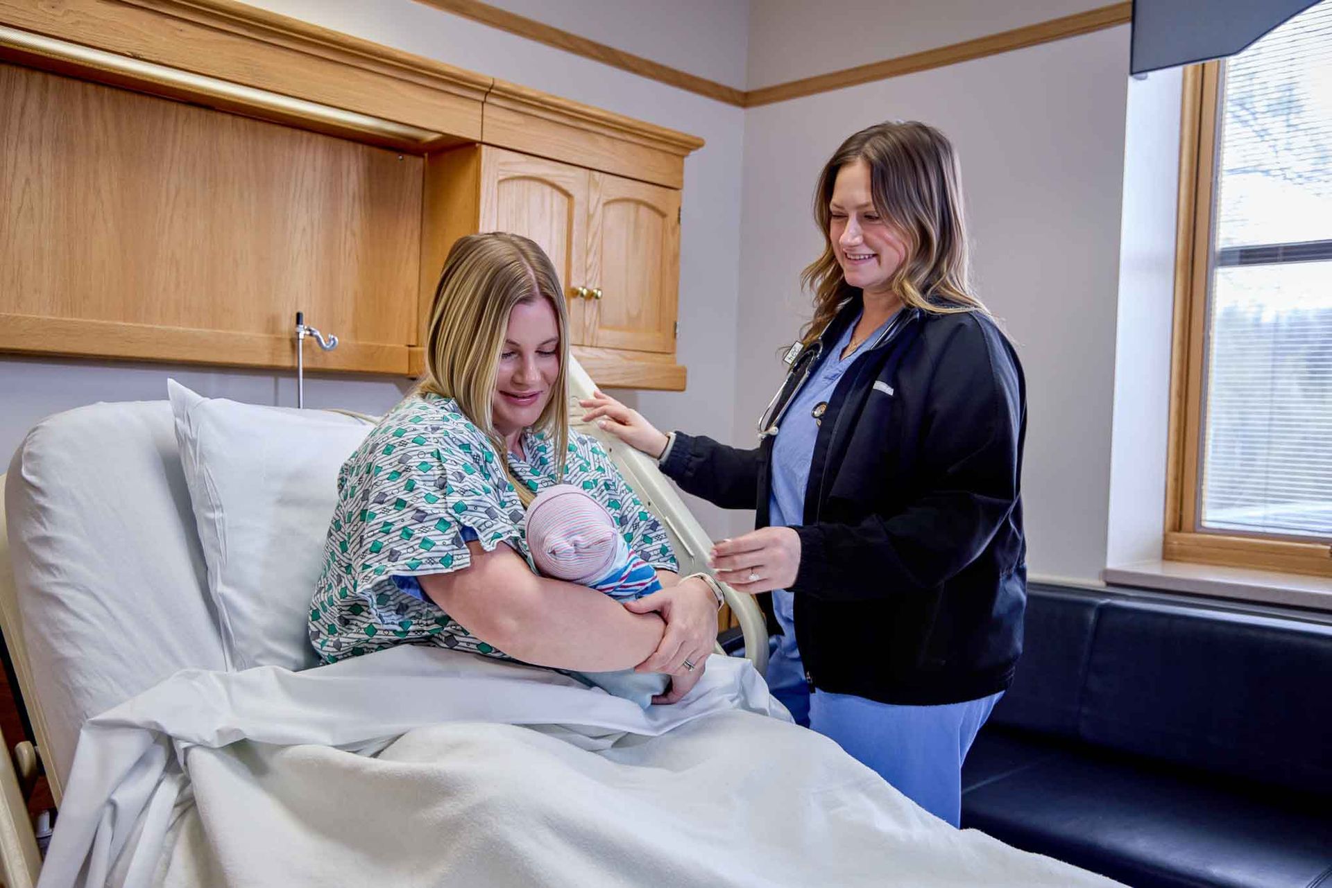 Woman in hospital bed holding baby, nurse standing beside her. They both smile. The room has wooden cabinets and a window.