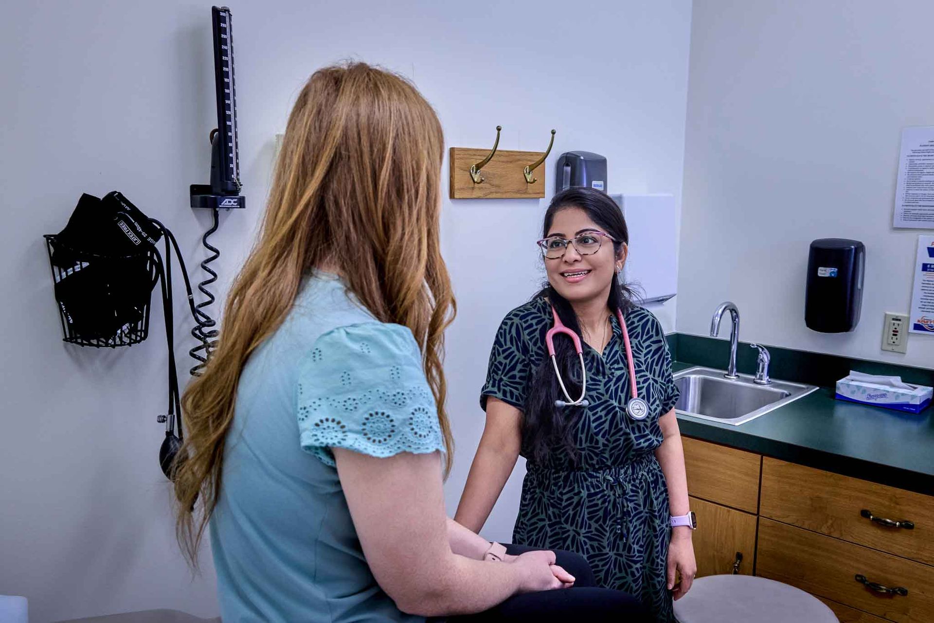 A doctor with a stethoscope smiles at a patient in an examination room.
