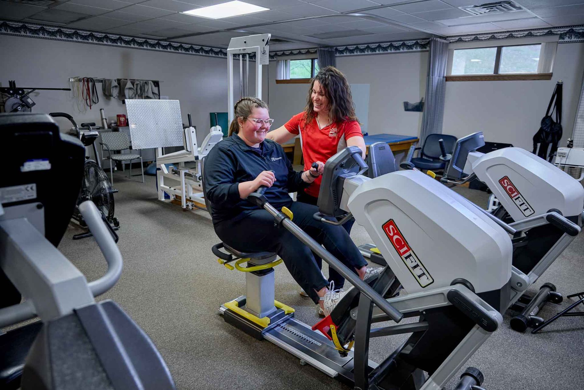 A woman smiles while using a seated leg exercise machine with assistance from a physical therapist in a gym.