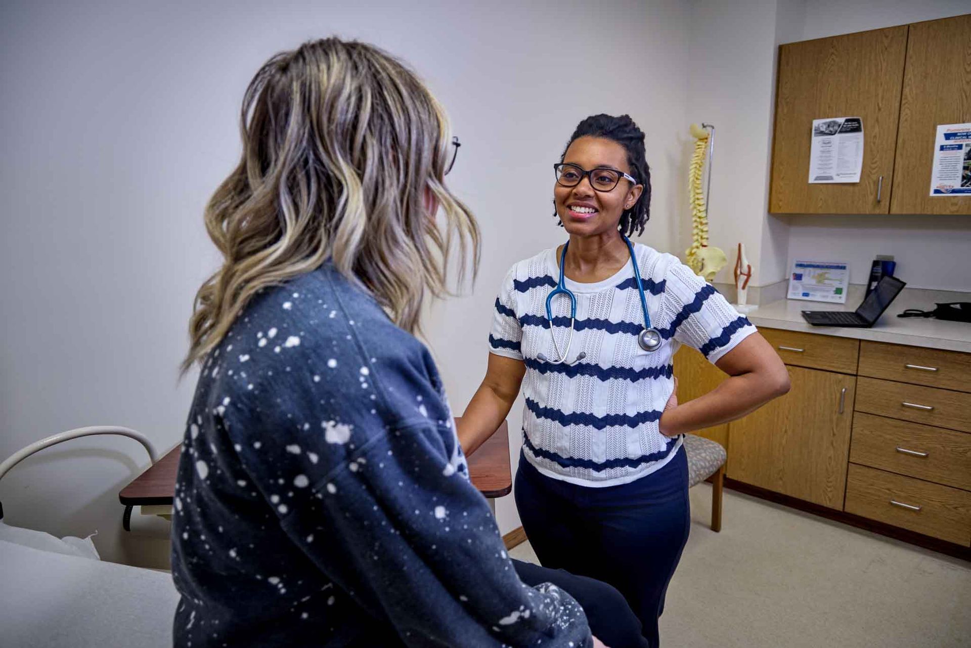 A doctor smiles at a patient in an exam room. The doctor, wearing glasses and a stethoscope.