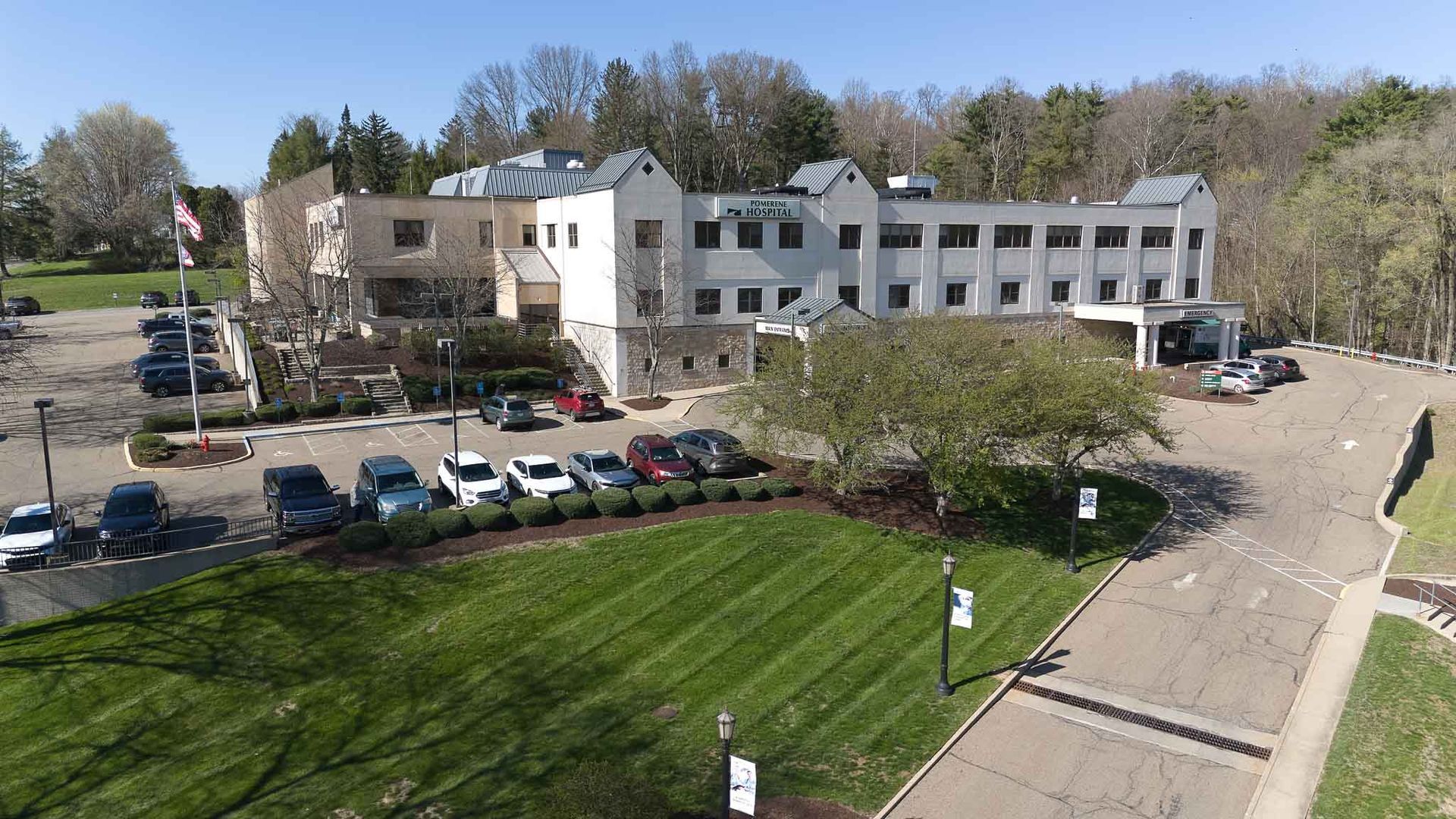 Aerial view of a light-colored castle-like building with parked cars, a grassy lawn, and surrounding trees under a sunny sky.