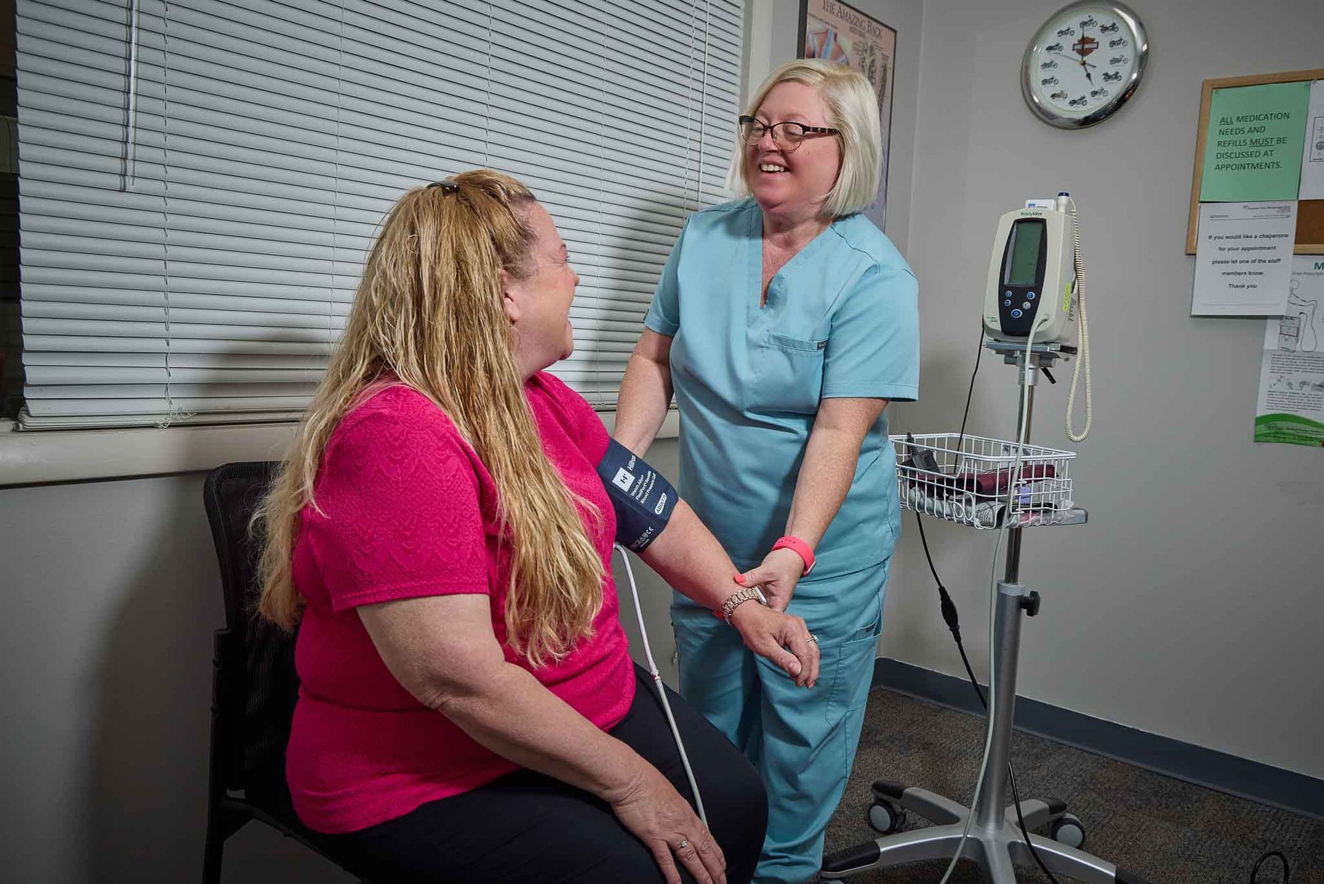 A healthcare worker takes a patient's blood pressure in an exam room. The worker smiles at the patient, who smiles back.