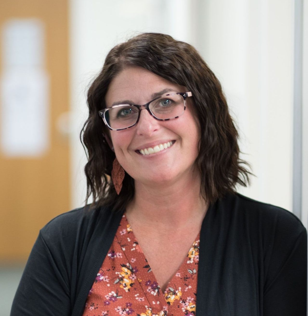 Smiling woman with glasses, brown hair, and a floral top, wearing a black cardigan, standing in a hallway.