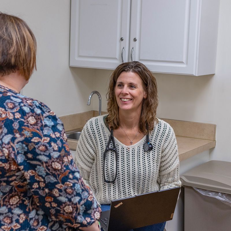 A smiling female doctor with a stethoscope, sitting with a patient in an exam room. She is holding a laptop.