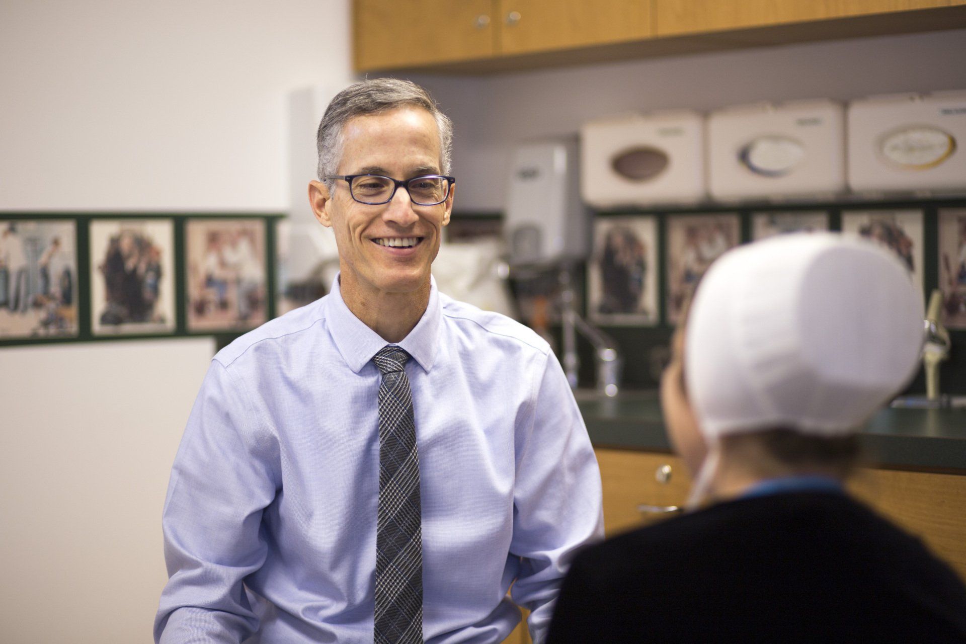 A smiling man in a blue shirt and glasses speaks to a person wearing a white head covering in a doctor's office.