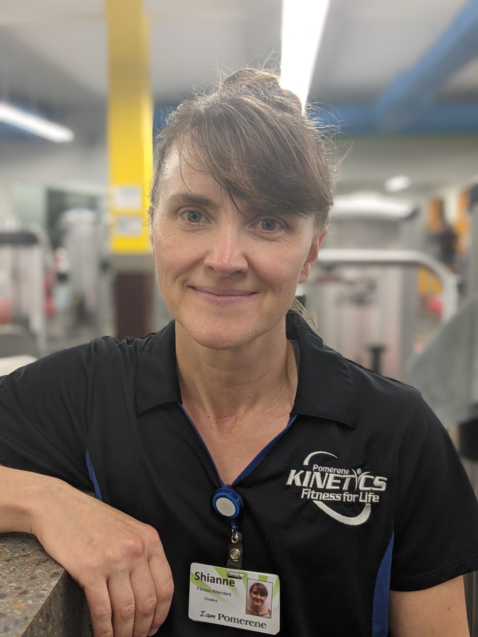 Woman with brown hair in a gym wearing a black Kinetic Fitness shirt with a name tag smiling at the camera.