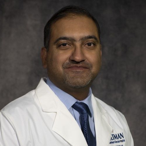 A smiling man with a beard wearing a white coat and tie, likely a doctor, against a gray background.