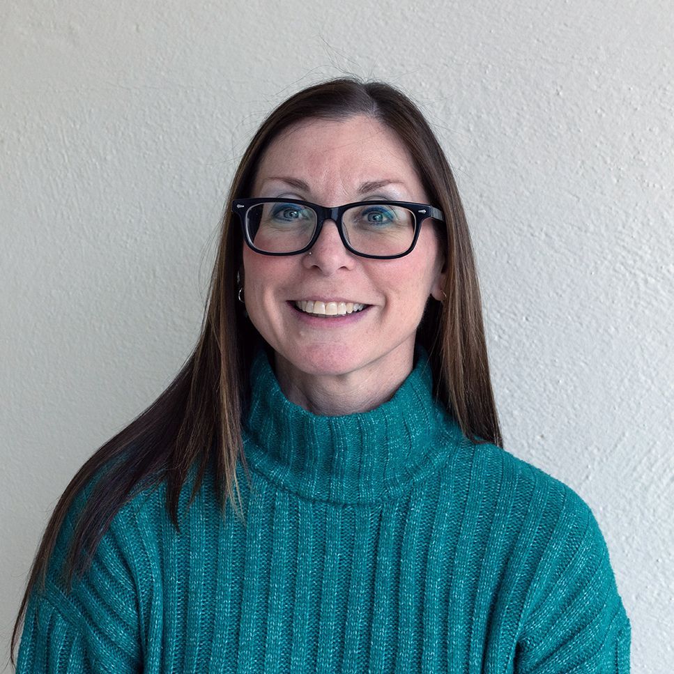 Woman with brown hair wearing glasses and a teal turtleneck sweater smiles in front of a beige wall.